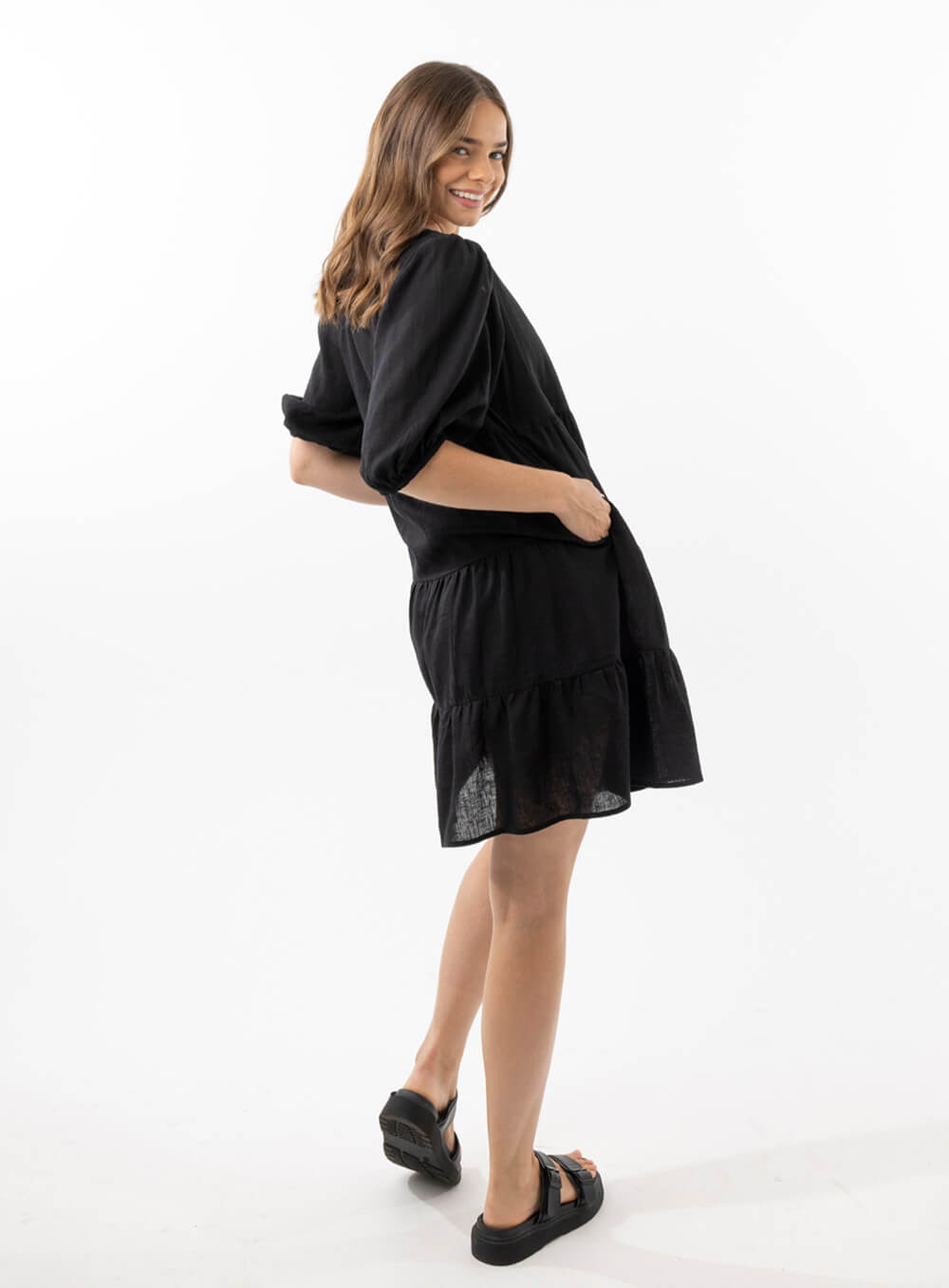 The Eleanor mini dress in black features a flowing layered a line shape with draping linen/viscose fabric, puff 3/4 sleeves with elastic cuff which can be pushed up to the elbow, a dropped v shape neckline with a square finish.