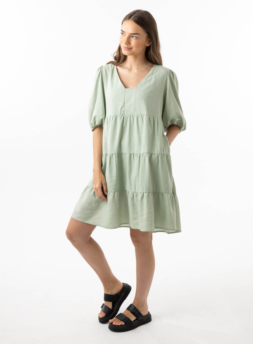 The Eleanor mini dress in sage features a flowing layered a line shape with draping linen/viscose fabric, puff 3/4 sleeves with elastic cuff which can be pushed up to the elbow, a dropped v shape neckline with a square finish.