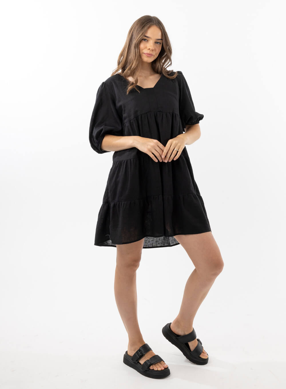 The Eleanor mini dress in black features a flowing layered a line shape with draping linen/viscose fabric, puff 3/4 sleeves with elastic cuff which can be pushed up to the elbow, a dropped v shape neckline with a square finish.